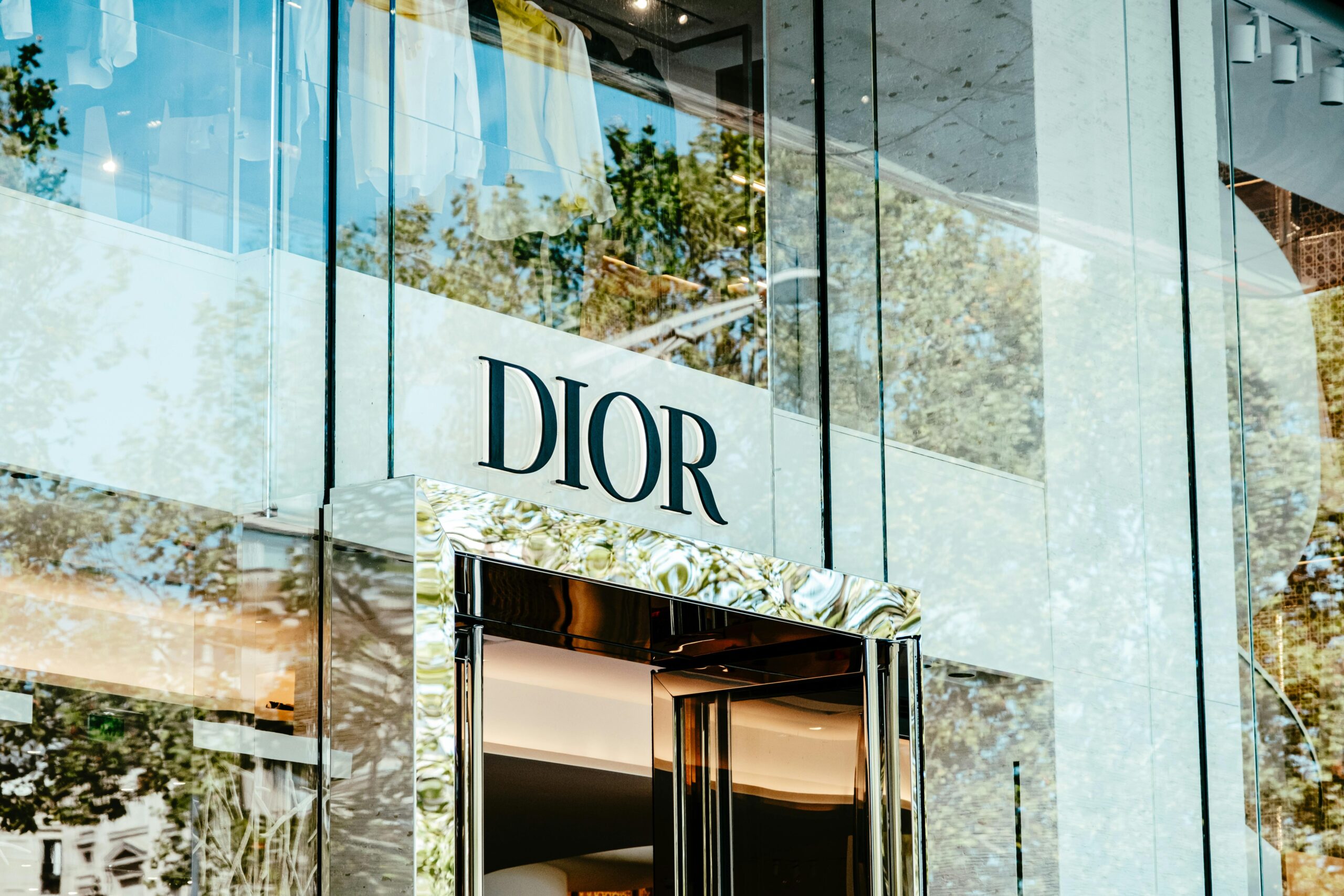 Exterior of a Dior store with glass facade reflecting trees and architecture.